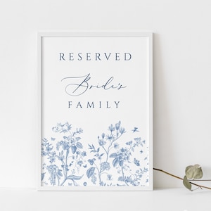 May include: A framed white sign with the words "RESERVED Bride's FAMILY" in navy blue. The bottom of the sign features a blue floral design. A sprig of greenery is in the lower right corner.