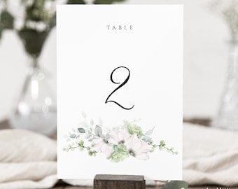 May include: White table number card with green foliage and white flowers. The card says "TABLE 2" in black text.