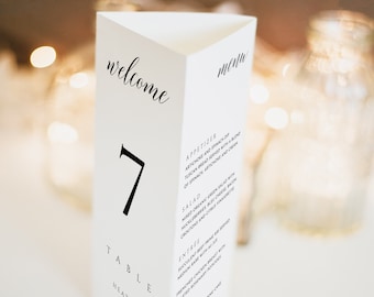 May include: A white triangular table number card with the number "7" in black font. The card also includes the text "welcome" and "menu" in black cursive font. The text "TABLE" is in black font, followed by the names "HEATHER & MICHAEL" in black font. The card also includes a sample menu with the headings "APPETIZER", "SALAD", "ENTREE", and "DESSERT".