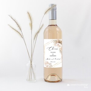 May include: A white wine bottle with a label featuring a gold geometric design and a floral illustration. The label reads "Cheers to Love & Happiness" and includes the names "Anika and Michael" and the date "May 23, 2023".