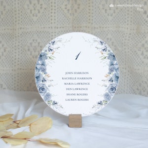 May include: A round, white table number card with a blue floral design. The number "1" is printed in black at the top. The names "John Harrison", "Rachelle Harrison", "Maria Lawrence", "Den Lawrence", "Shane Rogers", and "Lauren Rogers" are printed in black below the number.