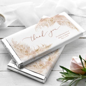 Puede incluir: Dos envoltorios de barra de chocolate blanco con un mensaje de agradecimiento y un diseño de acuarela de hierba de la pampa. El texto dice "¡Gracias por celebrar con nosotros! Alisha & Sabastian 12 de mayo de 2023".