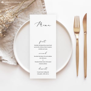 May include: A white menu card with black text listing the menu items for a wedding reception. The menu includes artichoke and spinach dip, beef prime rib, French chicken breast, and coconut layer wedding cake. The card is on a white plate with gold silverware.