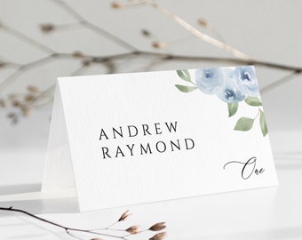 May include: A white place card with blue watercolour flowers in the top right corner. The text "ANDREW RAYMOND" is printed in black in the centre of the card. The number "One" is written in cursive in the bottom right corner.