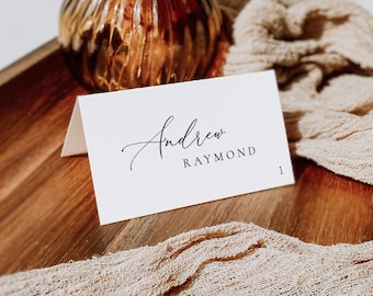 May include: White place card with black calligraphy that reads "Andrew Raymond" and the number "1" printed in the bottom right corner.