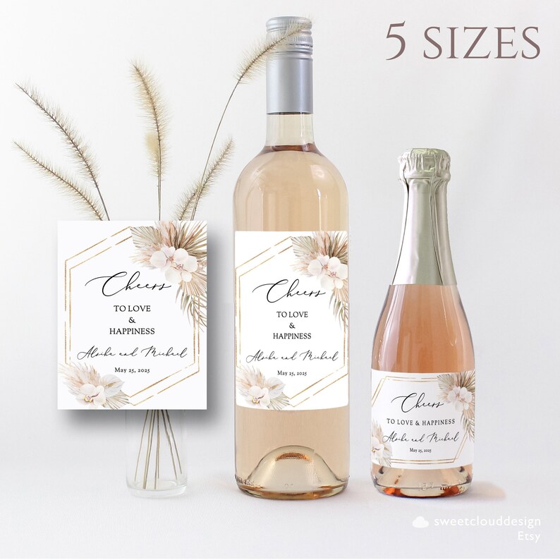 May include: Wedding wine bottle labels with a boho chic design featuring pampas grass, white flowers, and gold geometric shapes. The labels say "Cheers to love and happiness" with the couple's names and wedding date.