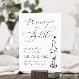 May include: A white sign with black text that reads "Message in a Bottle".  The sign also includes a black and white illustration of a bottle with a rolled up note inside.  The text below the illustration reads "Please leave a note for the Mr. and Mrs. to read on their first anniversary."