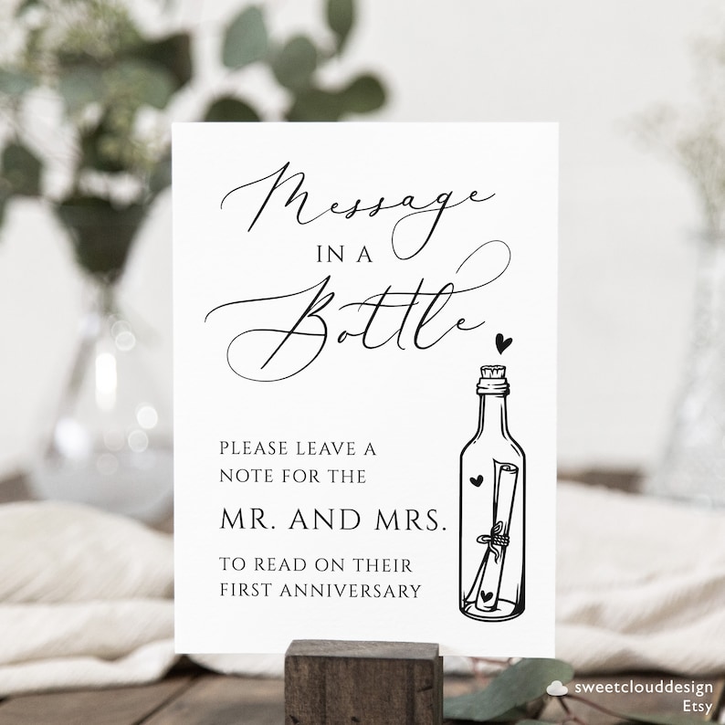 May include: A white sign with black text that reads "Message in a Bottle".  The sign also includes a black and white illustration of a bottle with a rolled up note inside.  The text below the illustration reads "Please leave a note for the Mr. and Mrs. to read on their first anniversary."