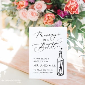 May include: A white sign with black text that reads "Message in a Bottle".  The sign also has a black and white illustration of a bottle with a rolled up note inside.  The sign says "Please leave a note for the Mr. and Mrs. to read on their first anniversary."