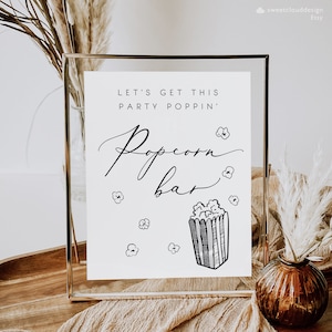 May include: A white sign with a gold frame that says "Let's get this party poppin' Popcorn bar" in black script. The sign has a black and white illustration of a popcorn bucket with popcorn spilling out.