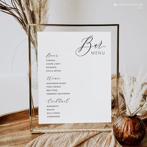 May include: A white framed sign with a black calligraphy font that reads "Bar Menu". The sign lists a variety of beer, wine, and cocktail options. The sign is sitting on a wooden surface with a small brown vase and dried flowers.