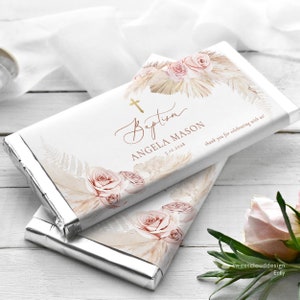 May include: A white chocolate bar wrapper with a gold cross, the words "Baptism" and "Angela Mason", and the date "5.12.2018". The wrapper features a watercolor design of pink roses, dried palm leaves, and a gold glitter background.
