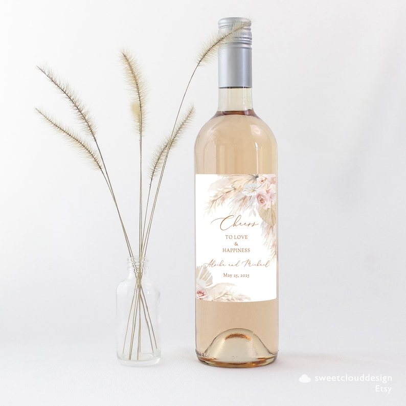 Puede incluir: Una botella de vino blanco con una etiqueta personalizada que presenta un dise&ntilde;o boho con hierba de la pampa seca y flores. La etiqueta dice "Cheers to love and happiness" con los nombres "Alexa y Michael" y la fecha "May 21, 2025".