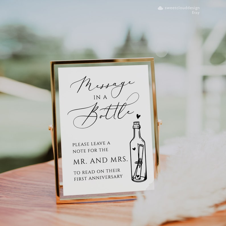 May include: A white sign in a gold frame with the text "Message in a Bottle" and a black and white illustration of a bottle with a rolled up note inside. The sign says "Please leave a note for the Mr. and Mrs. to read on their first anniversary."