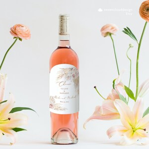Puede incluir: Una botella de vino rosado con una etiqueta blanca que presenta un dise&ntilde;o floral y el texto "Cheers to love and happiness" con los nombres "Alexa y Michael" y la fecha "May 20, 2022".
