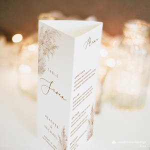 May include: A white table number card with a brown floral design and the text "Table Seven". The card also includes the names "Heather & Michael" and the date "August 12, 2011". The card is decorated with a watercolour-style illustration of pampas grass.