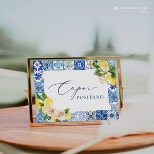 May include: A white sign with a blue and yellow border featuring a watercolor design of lemons and flowers. The sign says "Capri Positano" in a cursive font.