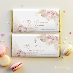 Può includere: Due involucri di barretta di cioccolato bianco con bordi dorati. Gli involucri presentano un design ad acquerello di rose rosa, erba della pampa e vegetazione. Il testo sugli involucri recita "Holy Communion" e "Angela Mason" con la data "5.12.2028".