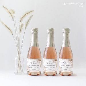 May include: Three mini champagne bottles with gold foil labels. The labels have a geometric design with a floral pattern and the text "Cheers to love and happiness" with a name and date.