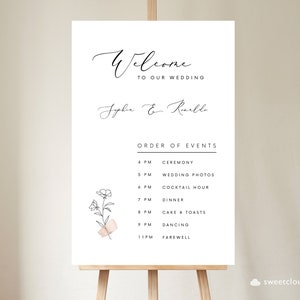 May include: A white wedding sign with black calligraphy that reads "Welcome to our wedding, Sophie & Raille". The sign also includes a black and white illustration of a flower with a pink ribbon and a list of wedding events with times.