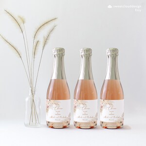 Puede incluir: Tres mini botellas de champ&aacute;n con etiquetas personalizadas. Las etiquetas presentan un dise&ntilde;o floral de acuarela con una fuente de escritura dorada que dice "Cheers to love and happiness" y los nombres "Alora y Michael".