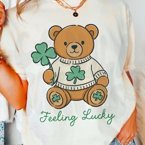 May include: Cream-colored t-shirt featuring a brown teddy bear holding a green shamrock. The bear wears a white sweater with a shamrock design. The words "Feeling Lucky" are printed in green script below the bear.