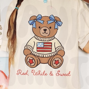 K&ouml;nnte beinhalten: Cremefarbenes T-Shirt mit einem braunen Teddyb&auml;ren, der einen Pullover mit amerikanischer Flagge und blauen Schleifen tr&auml;gt. Der Text "Red, White & Sweet" ist darunter gedruckt. Das Shirt ist ein l&auml;ssiger, bequemer Stil.