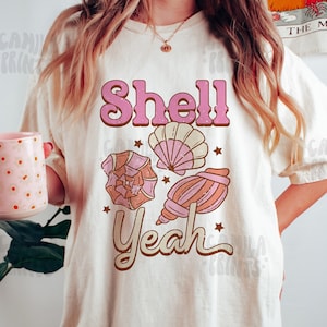 Pode incluir: Uma camiseta branca com o texto "Shell Yeah" em letras rosa e marrom. A camiseta apresenta um gráfico de quatro conchas em rosa e marrom.