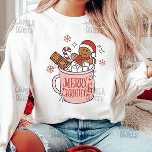 May include: White sweatshirt with a pink mug design featuring a gingerbread man, candy cane, marshmallows, and chocolate. The mug has the text "Merry & Bright" on it.