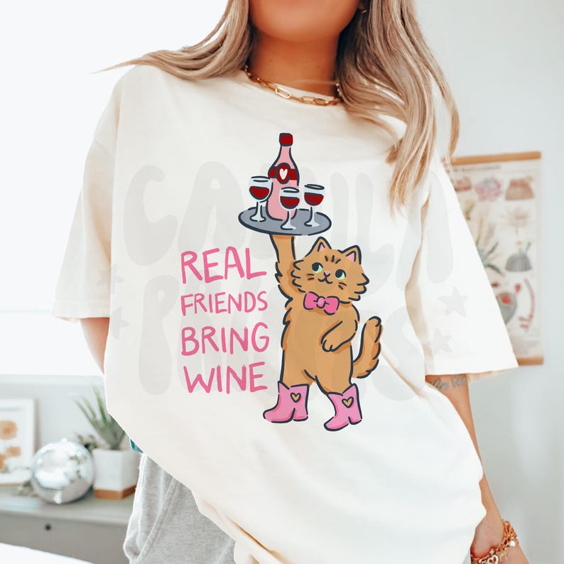 May include: Cream-colored t-shirt with a graphic of a cat holding a tray with wine glasses and a bottle. The text "REAL FRIENDS BRING WINE" is printed in pink. The cat wears a pink bow tie and boots.