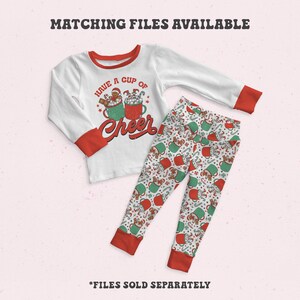 May include: White long-sleeved shirt with red trim and a graphic of two mugs of hot chocolate with marshmallows and a gingerbread man. The text "Have a cup of cheer" is printed on the shirt. White trousers with a red and green pattern of mugs of hot chocolate.