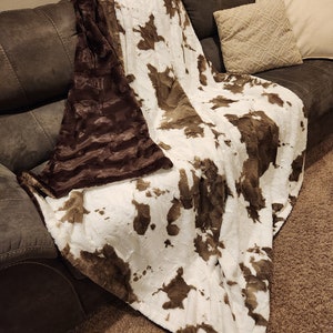 May include: A brown and white faux fur throw blanket with a cowhide pattern. The blanket is draped over a brown sofa.