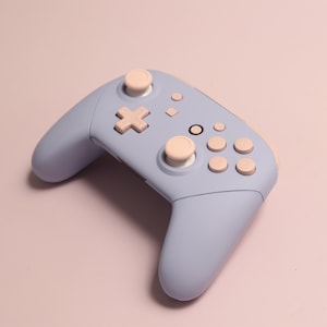 May include: A light blue video game controller with pink buttons. The controller has a directional pad, two analogue sticks, and several face buttons.