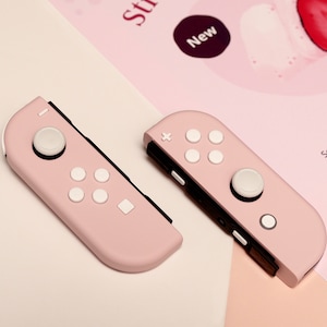 May include: Two pink Nintendo Switch Joy-Con controllers with white buttons. The controllers have a textured surface and are designed for gaming.