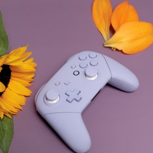 Könnte beinhalten: Ein lilafarbener Videospiel-Controller mit weißen Knöpfen. Der Controller liegt auf einer lilafarbenen Oberfläche mit einer gelben Sonnenblume und orangefarbenen Blütenblättern.