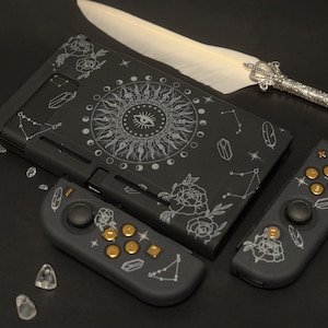 Może przedstawiać: Czarna konsola Nintendo Switch z niestandardowym wzorem przedstawiającym białą mandalę z okiem w środku, otoczoną księżycami i gwiazdami. Konsola jest również ozdobiona białymi różami i piórami. Obraz przedstawia konsolę i dwa czarne kontrolery ze złotymi przyciskami.