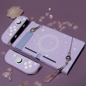 Puede incluir: Una consola Nintendo Switch lavanda con un diseño personalizado que presenta una luna blanca y estrellas en un círculo. La consola está rodeada de cristales rosas y un collar con un dije plateado.