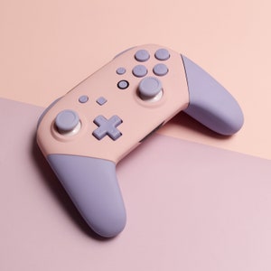 May include: A pastel pink and lilac wireless game controller. The controller has a pink body with lilac handles and buttons. It features a directional pad, analog sticks, and action buttons. The controller is designed for gaming.