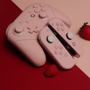 May include: A pink Nintendo Switch controller with two white buttons and a pink Joy-Con controller with two white buttons. The controllers are on a pink and red surface with a red strawberry.