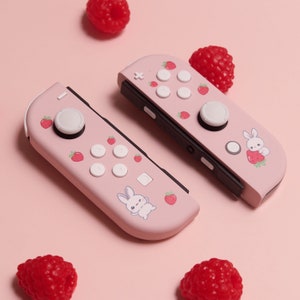 May include: Two pink Nintendo Switch Joy-Con controllers with a strawberry and bunny design. The controllers have white buttons and a pink background. The controllers are surrounded by pink raspberries.