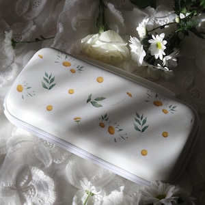 Puede incluir: Estuche rectangular blanco adornado con un diseño floral de margaritas. El estuche tiene una cremallera blanca y está decorado con margaritas y hojas verdes estilo acuarela. Fondo de tela blanca texturizada.