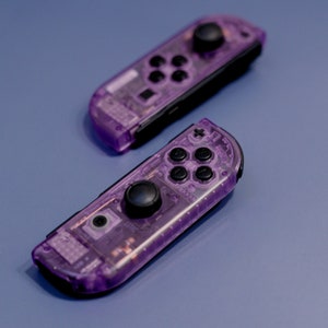 May include: Two purple translucent Nintendo Switch Joy-Con controllers with black buttons. The controllers have a clear, see-through design, revealing the internal components.