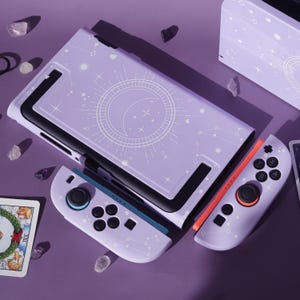 May include: A lilac gaming console with coordinating controllers and a protective case. The case and controllers have a white celestial design featuring moons, stars, and constellations. The console is on a purple surface with crystals and a tarot card.