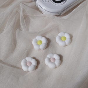 May include: Four white ceramic flower-shaped phone grips with yellow and pink centres. The grips are on a white mesh surface.