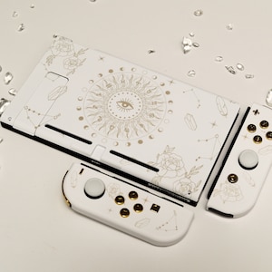 May include: White Nintendo Switch console with a custom design featuring a gold sun with an eye, constellations, and flowers. The console is separated into three pieces: the main console, the left joy-con, and the right joy-con.