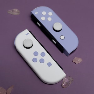 May include: Two Nintendo Switch controllers, one white and one lavender. The white controller has a white button and four purple buttons. The lavender controller has a white button and four white buttons.