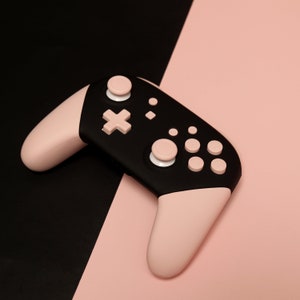 Puede incluir: Un mando de videojuegos negro y rosa con varios botones. El mando está sobre una superficie negra y rosa.