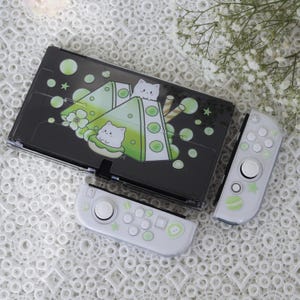 Op de afbeelding: Een witte Nintendo Switch-console met een aangepaste skin met een groen en wit ontwerp met cartoonkatten en bubble tea. De console wordt getoond met twee witte Joy-Con-controllers.