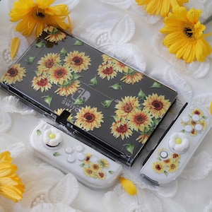Peut inclure: Une console Nintendo Switch noire avec un étui transparent à motif de tournesols. La console est accompagnée de deux manettes Joy-Con blanches avec des motifs de tournesols et des boutons en forme de marguerite blanche.