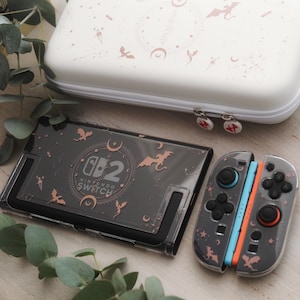 Peut inclure: Ensemble d'accessoires pour console Nintendo Switch. La console et les manettes sont gris foncé avec des accents or rose, notamment des chauves-souris, des lunes et le logo Nintendo Switch. Une housse de transport blanche avec des motifs assortis est également incluse.